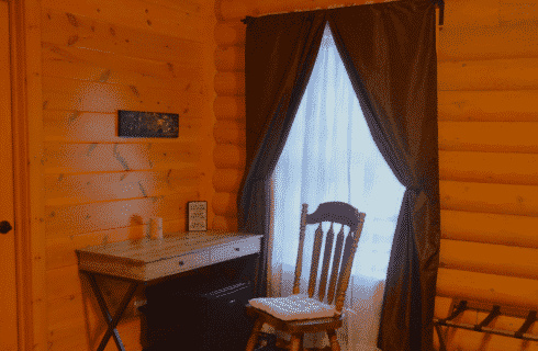 Corner of a bedroom in a log home with writing desk and chair near a window with white and brown curtains