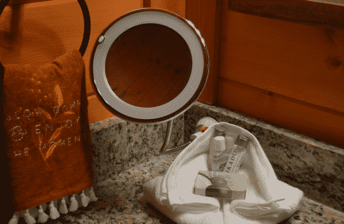 Corner of a bathroom counter with hanging towel, mirror and toiletries nestled in a white towel