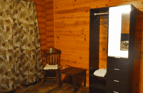 Corner of a log home bedroom with sitting chair, table, closet and curtains