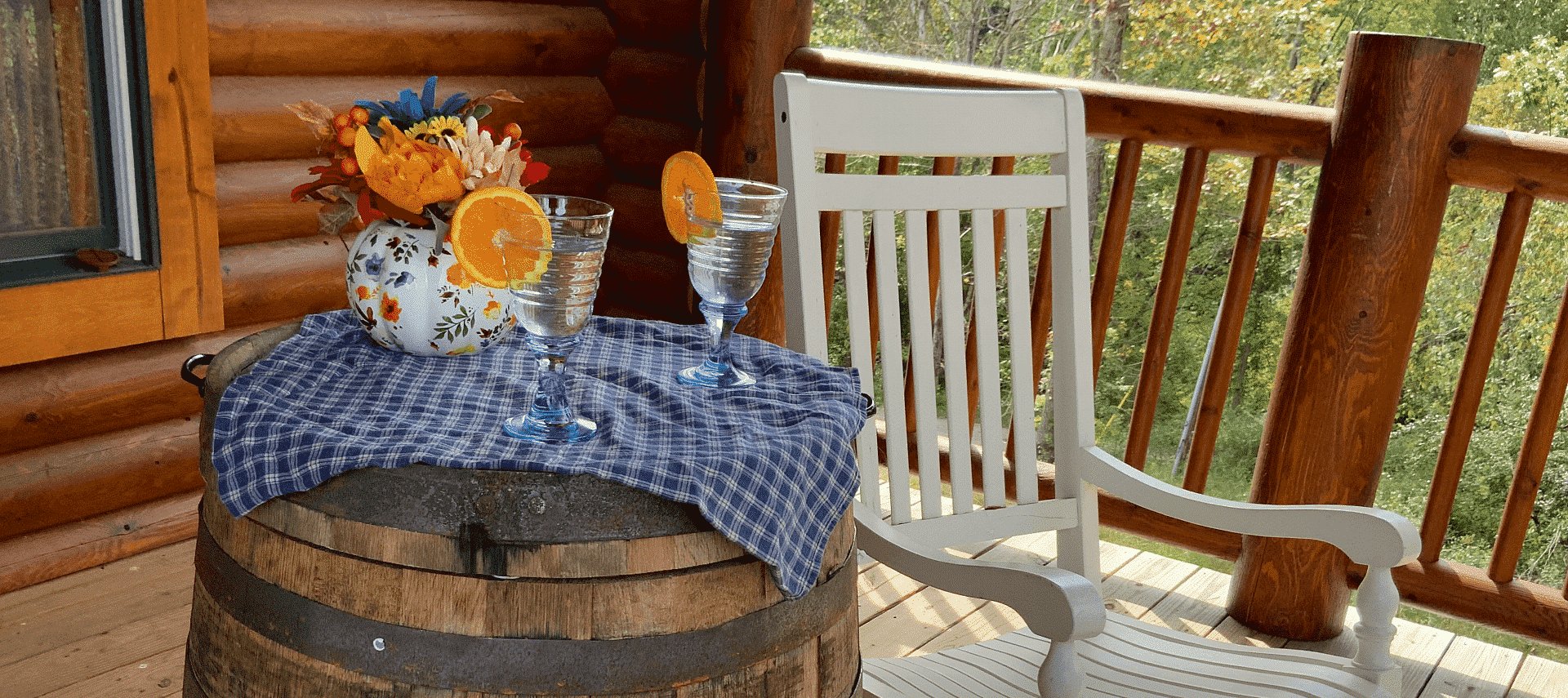 Outside porch of log home with white rocking chair next to a barrel holding glasses of water on a blue checkered cloth