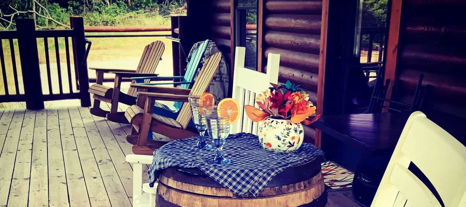 Front porch of a log home with adirondack chairs, white rocking chair and barrel with glasses of water and vase of flowers