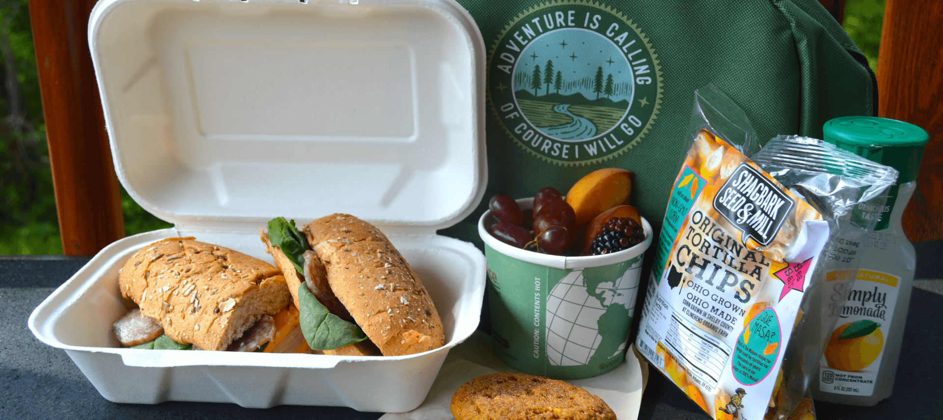 White container with sandwich, cup of fruit, bag of chips, lemonade and green carrying bag