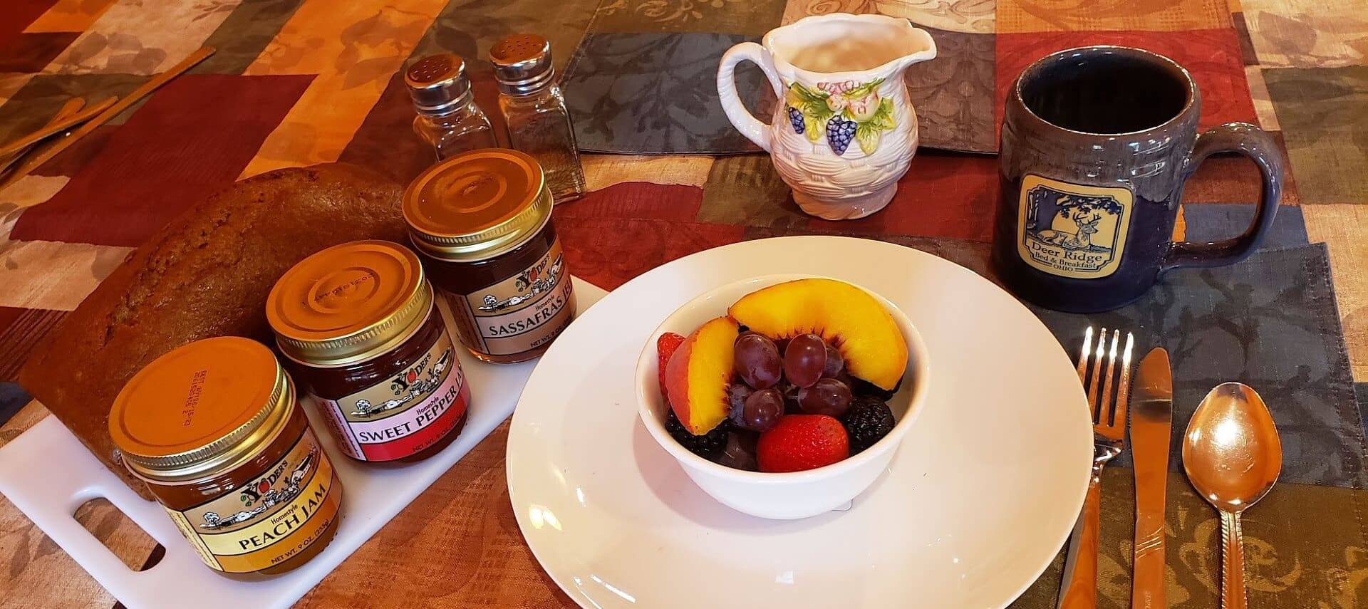 White round plate with a bowl of fruit next to jars of jam, loaf of bread and ceramic coffee mug