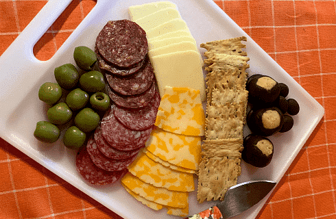 Square white plate with green olives, meat, cheeses, crackers and a knife