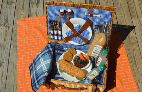 Open picnic basket with plate of sandwich, cookies and fruit and two lemonade bottles with blue checkered napkin